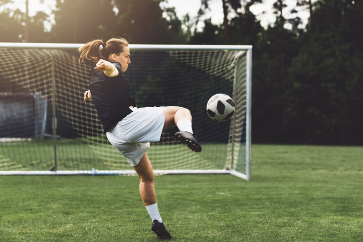 Women's soccer player kicking a ball into a net