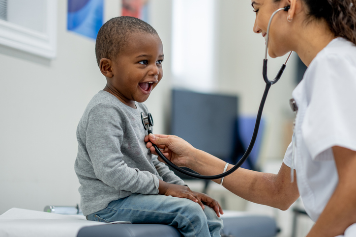 A young boy of African decent, sits up on an exam table as a female Pediatrician preforms a check-up on him.