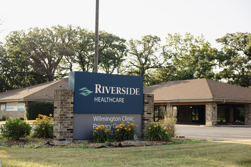 Riverside Healthcare Wilmington Primary Care sign stands prominently in front of the brick clinic on a bright sunny day.