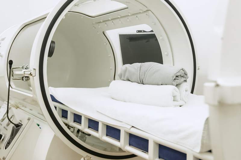 Interior view of a hyperbaric oxygen therapy chamber showing a comfortable patient bed with white linens and pillows, a viewing window, and the clear cylindrical treatment space at Riverside Healthcare's Wound Care Center