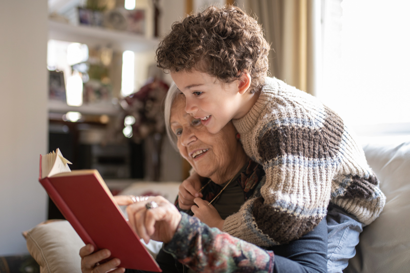 Grandmother sitting on a couch reading a book while her young grandson in a cozy striped sweater leans over her shoulder, both smiling in a warm living room.