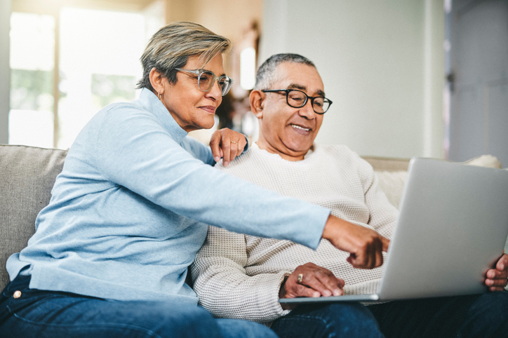 An older couple sits together on a sofa at home, smiling as the woman points to information on a laptop they're reviewing together.